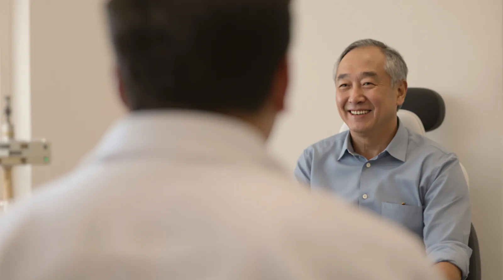 An older man smiles while talking with a physician seen from behind in a bright exam room